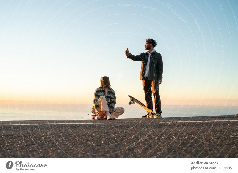 Glückliches multiethnisches Paar mit Skateboards, das gemeinsam Spaß auf der Straße hat Lächeln Sonnenuntergang Zusammensein Daumen hoch trampen gestikulieren