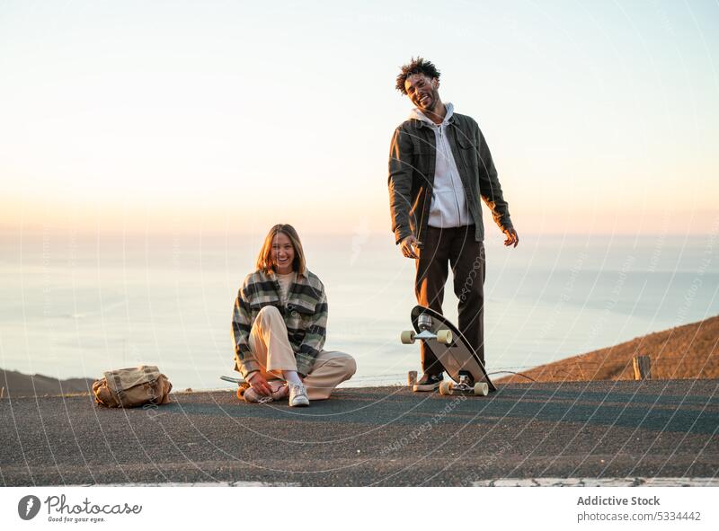 Glückliches multiethnisches Paar mit Skateboards, das gemeinsam Spaß auf der Straße hat Lächeln Sonnenuntergang Zusammensein Partnerschaft Liebe Skater heiter