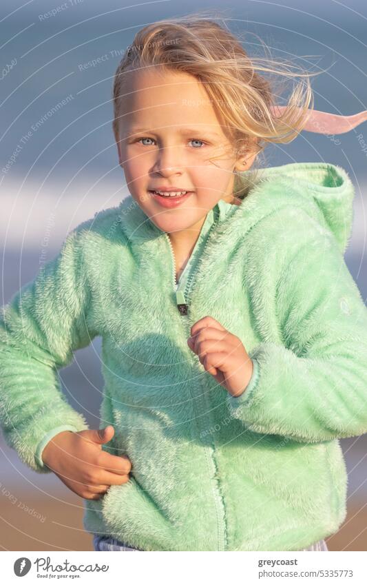 Ein 5-jähriges Mädchen in einer grünen Jacke läuft Kind Kindheit blond 5 Jahre alt Strand MEER Sand Meer Küste vertikal jung Auge blau Feiertag Lifestyle Himmel