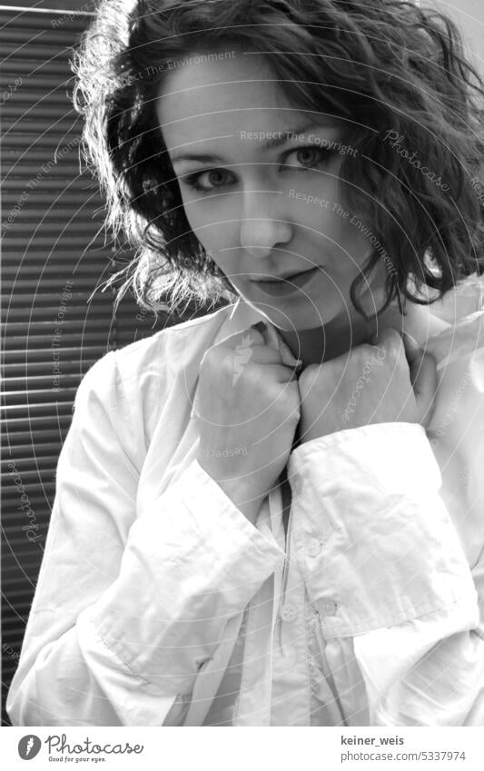 Eine Frau mit gelockten Haaren steht im weißen Hemd mit den Händen am Hemdkragen vor einer Jalousie am Fenster als Porträt in schwarzweiß Haarlocken Erwachsene