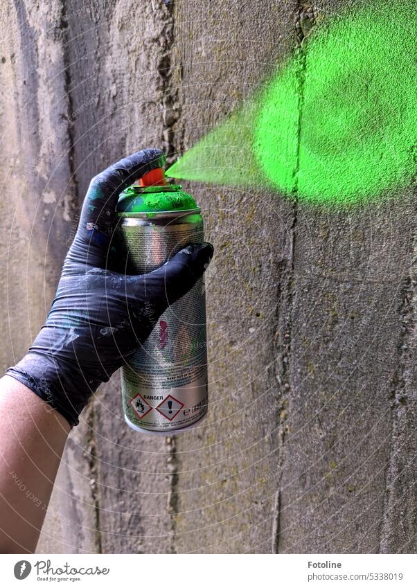Eine Hand, geschützt durch einen schwarzen Einmalhandschuh, drückt den Knopf einer Spraydose runter und sprüht eine graue Wand knallig grün an. Etwas farblosem einfach mal einen neuen Farbton geben!.