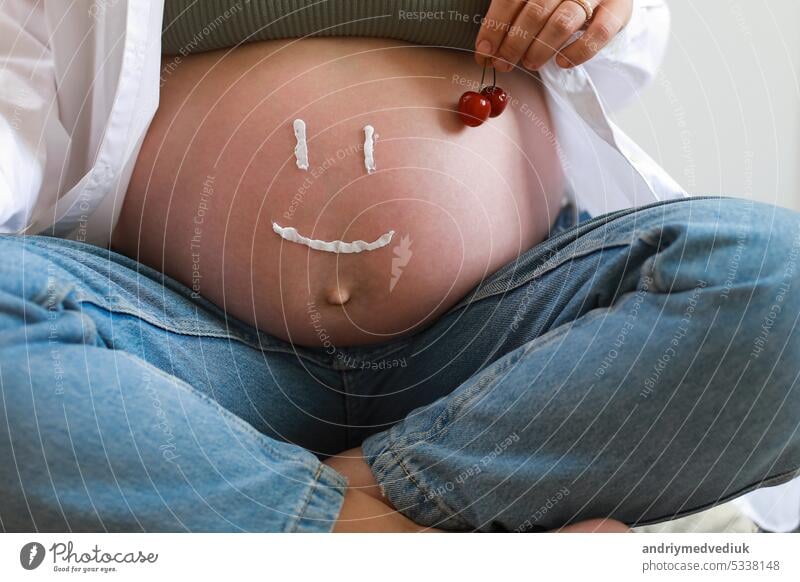 Nahaufnahme des Oberkörpers eines jungen schwangeren Models, das Feuchtigkeitscreme auf ihren Bauch aufträgt, um Dehnungsstreifen zu vermeiden. Zukünftige Mutter mit lustigem Lächeln von Feuchtigkeitscreme auf ihrem Bauch. Konzept für die Hautpflege in der Schwangerschaft