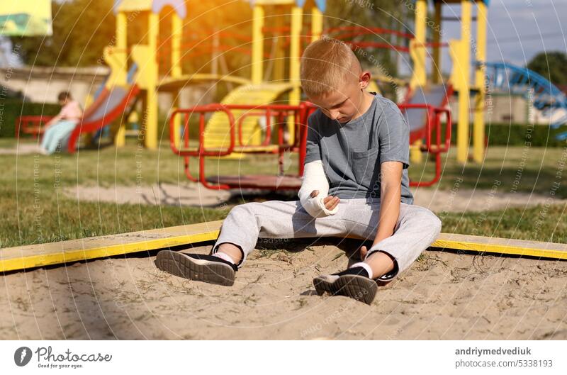 weinendes Kind mit gebrochenen Gliedmaßen Arm sitzt im Freien und schauen, wie Kind Mädchen spielt auf dem Spielplatz. Konzept der Gesundheit Unfall, und medizinische. die schlimmsten Sommerferien.