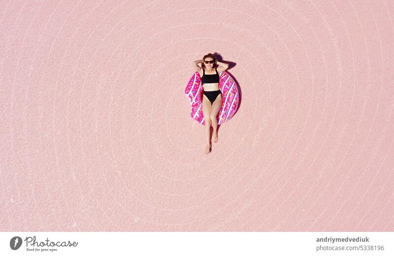Welttourismustag. Blick von oben auf eine junge glückliche Frau mit Sonnenbrille und schwarzem Bikini, die sich auf einem aufblasbaren rosa Donut in einem sonnigen rosa See sonnt. Sie genießt ihren Urlaub. Platz für Werbung