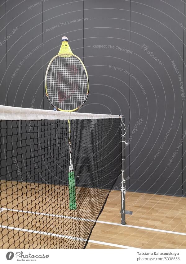 Ein Badmintonschläger hängt an einem Badmintonnetz, auf dem ein gelber Kunststoff-Federball auf einem Badmintonplatz liegt. im Innenbereich Sport Erholung Spiel