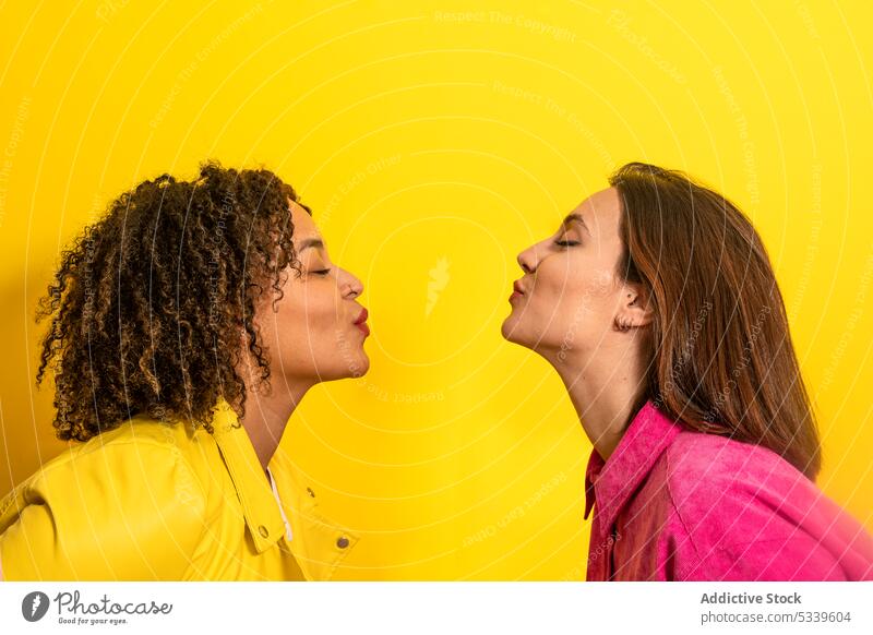 Glückliche multiethnische Freunde, die sich auf gelbem Hintergrund küssen Frauen Zusammensein heiter Lächeln Liebe Luftkuss Partnerschaft Freundin jung
