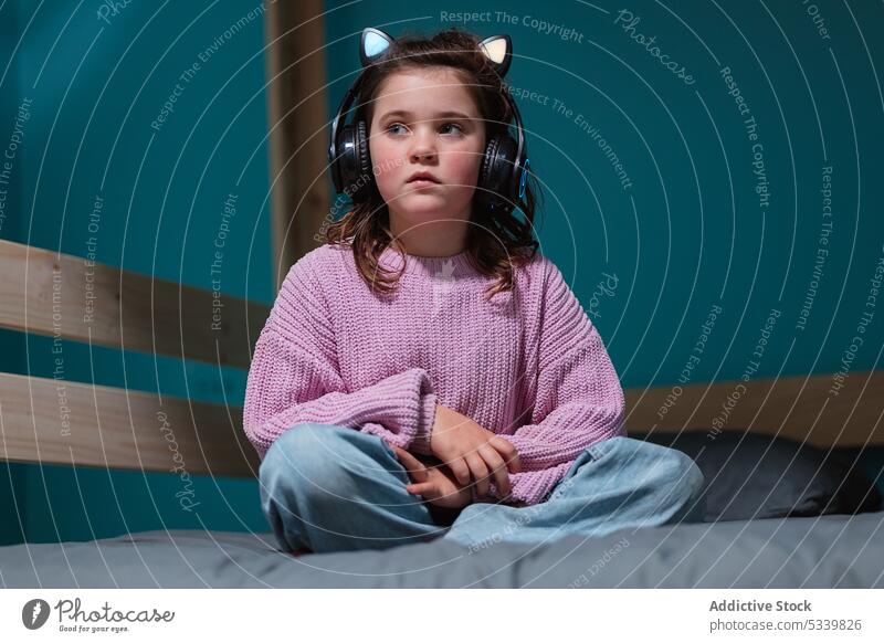 Ruhiges Kind mit Kopfhörern auf dem Bett sitzend Musik zuhören Beine gekreuzt bezaubernd nachdenklich Schlafzimmer Apparatur wenig heimwärts Gerät Komfort
