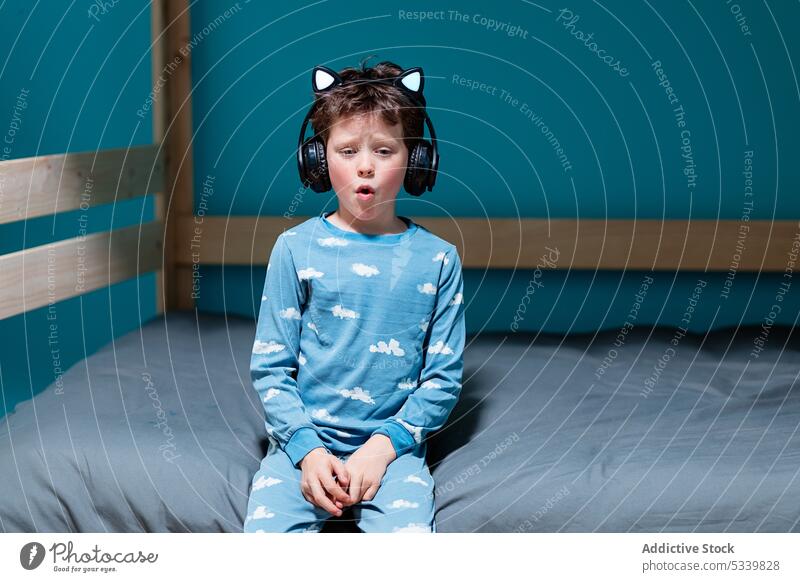 Niedliches Kind mit Kopfhörern auf dem Bett sitzend singen Gesang Musik zuhören Nacht Drahtlos niedlich Schlafzimmer Apparatur Kindheit heimwärts Gerät Audio
