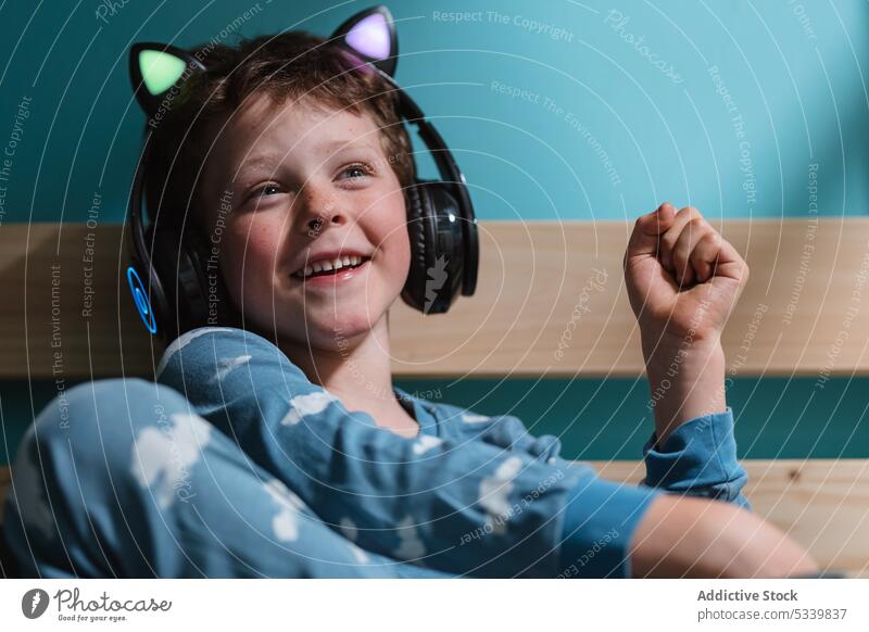 Niedliches Kind mit Kopfhörern auf dem Bett sitzend Musik zuhören Nacht Lächeln Drahtlos niedlich Schlafzimmer Apparatur Kindheit heiter heimwärts Glück Gerät