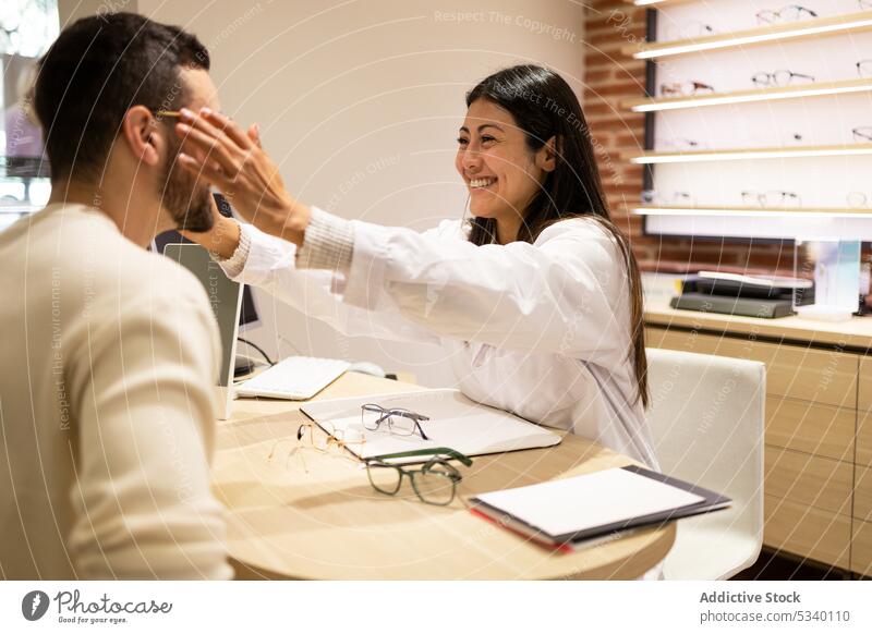 Fröhliche Augenärztin, die einem männlichen Kunden hilft, eine Brille zu tragen Mann wählen Laden optisch Lächeln Ophthalmologe Augenlicht Sehvermögen Werkstatt