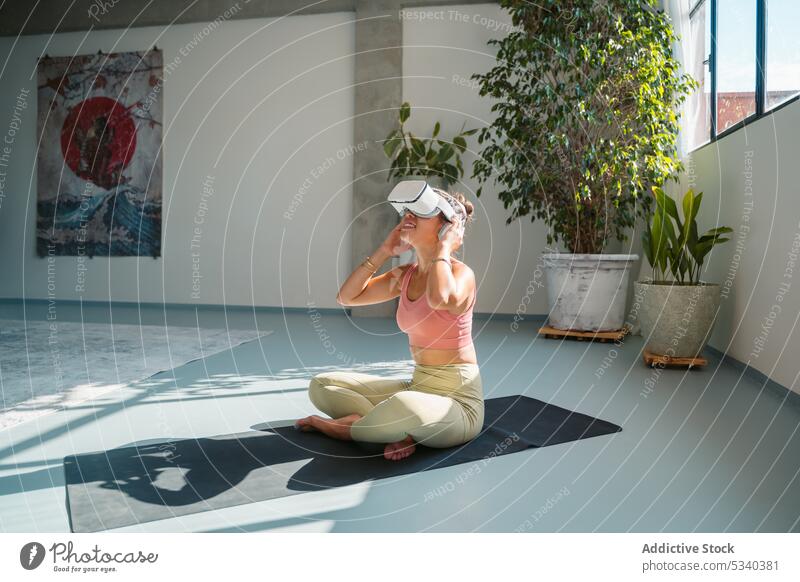 Lächelnde Frau in VR-Headset auf Yogamatte padmasana Lotus-Pose Virtuelle Realität Schutzbrille heiter Asana üben Wellness Barfuß meditieren Sportkleidung