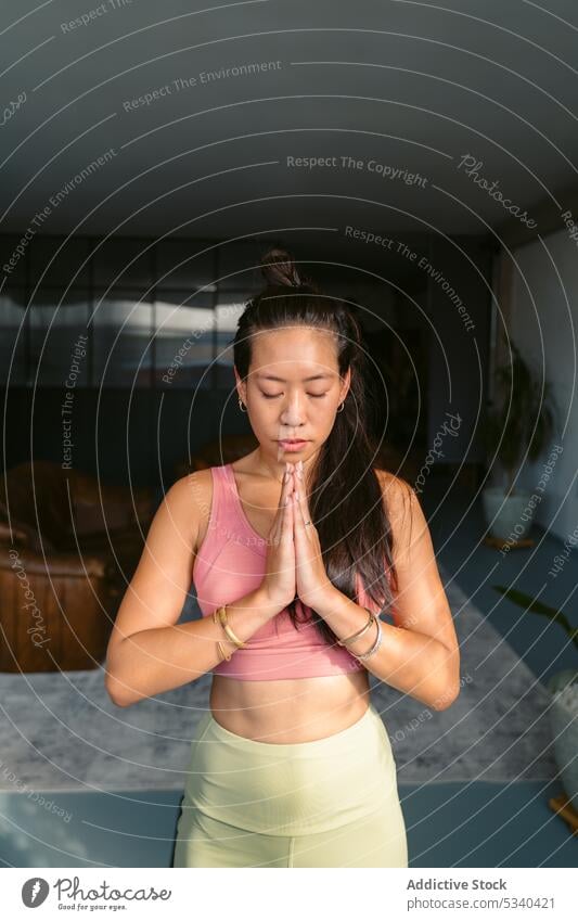 Ruhige asiatische Frau mit gefalteten Händen im Wohnzimmer Yoga Namaste meditieren heimwärts üben Achtsamkeit ruhig Augen geschlossen Asana Windstille Harmonie