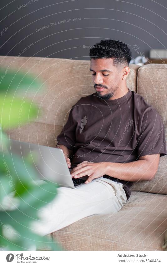 Ernster ethnischer Mann arbeitet zu Hause am Laptop Arbeit freiberuflich benutzend Tippen online abgelegen heimwärts Internet Konzentration jung Sofa