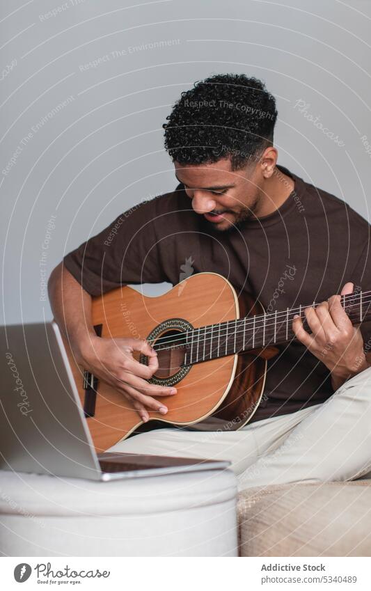Fröhlicher ethnischer Mann spielt Gitarre mit Laptop auf dem Sofa spielen benutzend positiv online Musik akustisch Gitarrenspieler Musiker Instrument Hobby jung