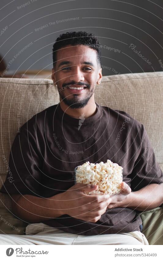 Glücklicher ethnischer Mann, der Popcorn isst und auf dem Sofa fernsieht zuschauen FERNSEHER essen heiter Wochenende Popkorn heimwärts Lächeln Afroamerikaner