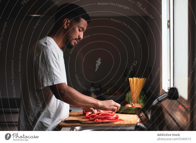 Fokussierter ethnischer Mann schneidet Paprika auf einem Schneidebrett Salatbeilage geschnitten Messer vorbereiten Gemüse Küche Koch Gesundheit Lebensmittel