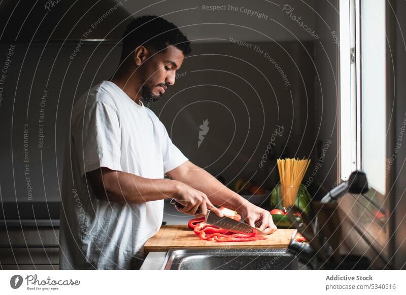 Fokussierter ethnischer Mann schneidet Paprika auf einem Schneidebrett Salatbeilage geschnitten Messer vorbereiten Gemüse Küche Koch Gesundheit Lebensmittel