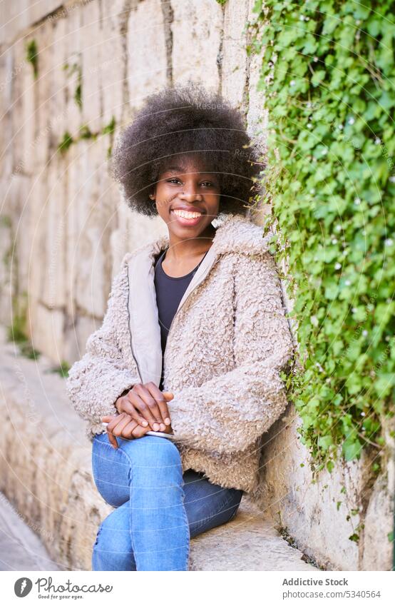 Fröhlicher schwarzer Teenager mit Afrofrisur sitzt auf einer Betonterrasse an einer Steinmauer Terrasse Mantel Glück urban positiv heiter Lächeln Freude