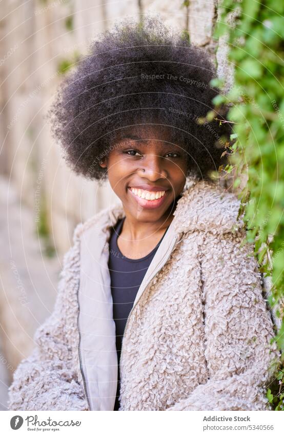 Fröhlicher schwarzer Teenager mit Afrofrisur auf Betonterrasse an Steinmauer Terrasse Mantel Glück urban positiv heiter Lächeln Freude Afroamerikaner Frau