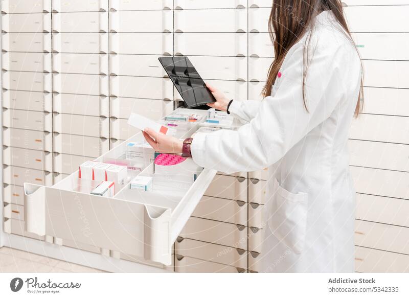 Crop Apotheker mit Tablette, während stehend durch geschlitzte Regale mit Medikamenten in modernen Speicher Wissenschaftler Labor Frau Drogerie Laden benutzend