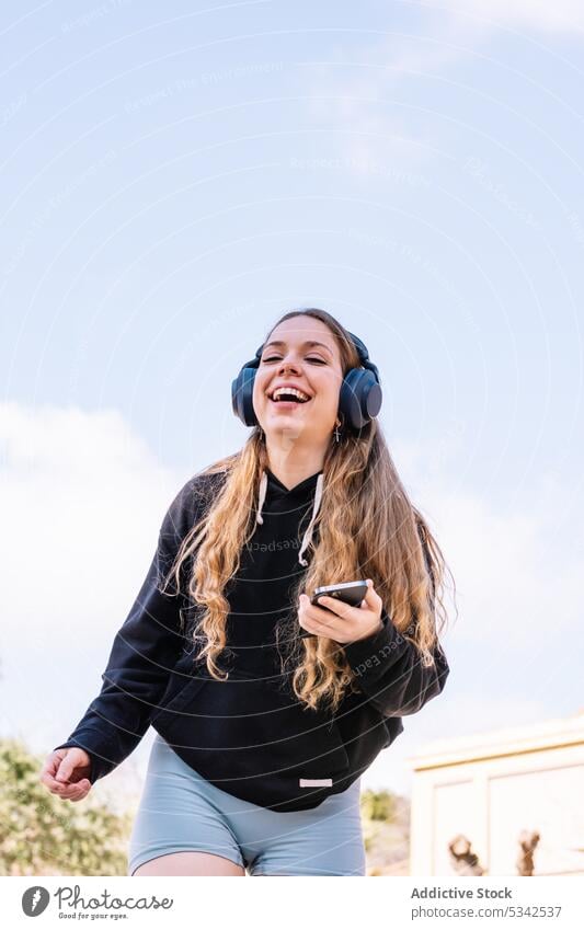 Lächelnde Frau mit Smartphone, die über Kopfhörer Musik hört zuhören benutzend heiter Freude genießen sorgenfrei Apparatur Drahtlos Glück Gerät jung lässig
