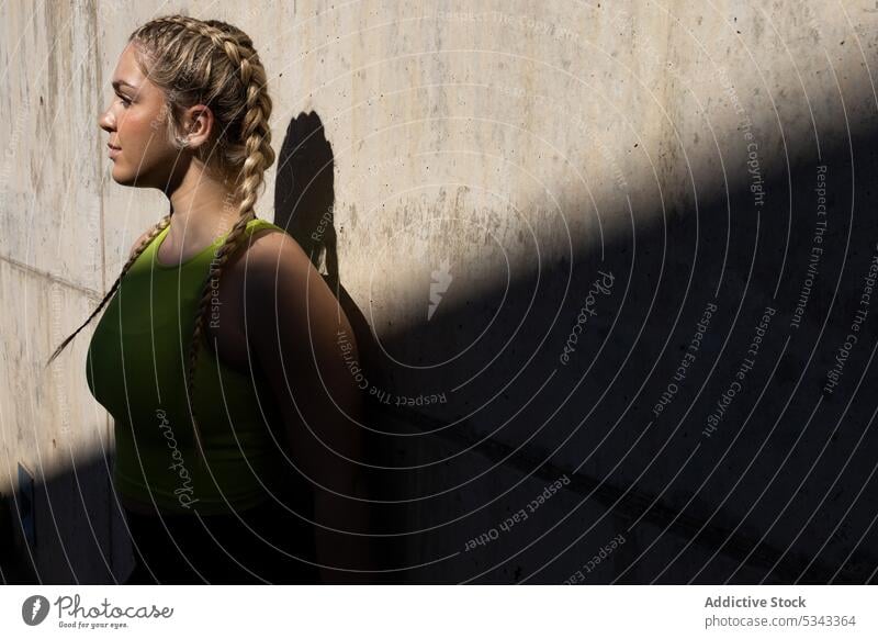 Junge Frau steht in der Nähe einer Betonwand mit Schatten Sportlerin Porträt selbstbewusst Athlet Sportbekleidung Wand Straße Sonnenlicht jung stehen verträumt