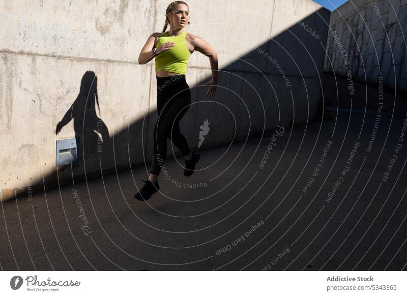 Fitte Sportlerin joggt auf asphaltierter Straße an einer Betonmauer Athlet Sportbekleidung joggen Schatten Asphalt laufen Training Wellness Frau jung Dame