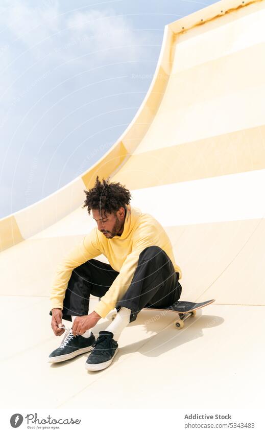 Stylish schwarzer Mann sitzt auf Skateboard über Skate-Rampe im Tageslicht Skater Skateplatz Schuhbänder Krawatte Turnschuh Aktivität Blauer Himmel Energie