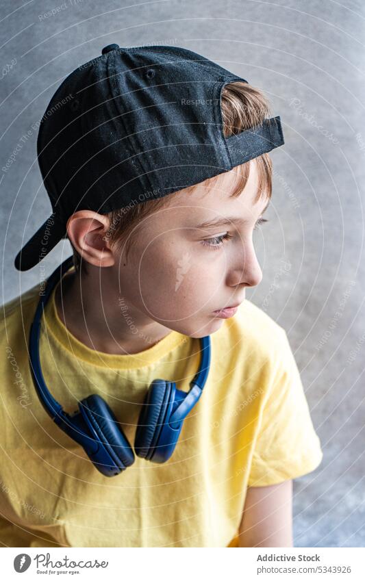 Seitenansicht eines nachdenklichen Jungen in gelbem T-Shirt und schwarzer Mütze mit Kopfhörern im Nacken, der vor einem unscharfen Hintergrund wegschaut schön