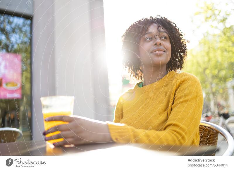 Glückliche schwarze Frau sitzt mit einem Glas Orangensaft Lächeln positiv orange Saft genießen froh angenehm Café heiter trinken jung krause Haare