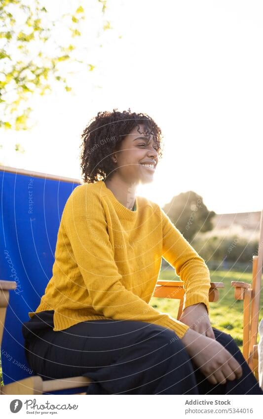 Fröhliche junge ethnische Frau sitzt auf einem Stuhl im Park Natur Baum Inhalt froh positiv Lächeln genießen Sommer Glück Hand auf die Wange Afroamerikaner