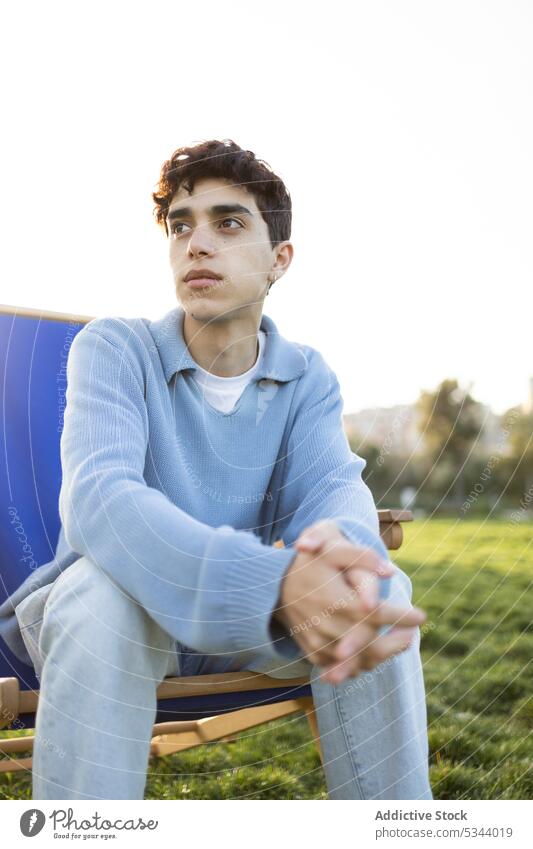 Nachdenklicher junger Mann sitzt auf einem Stuhl im Park sich[Akk] entspannen nachdenken Hände gefaltet Sommer besinnlich nachdenklich Windstille Sonnenlicht