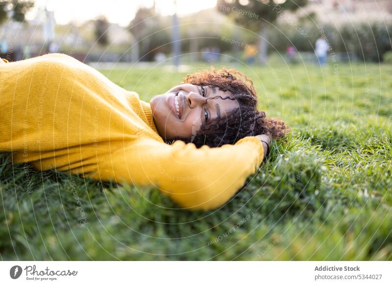 Glückliche ethnische Frau mit lockigem Haar im Gras liegend Lächeln Park Lügen Natur Baum Rasen grün Inhalt positiv jung Afroamerikaner schwarz Pullover