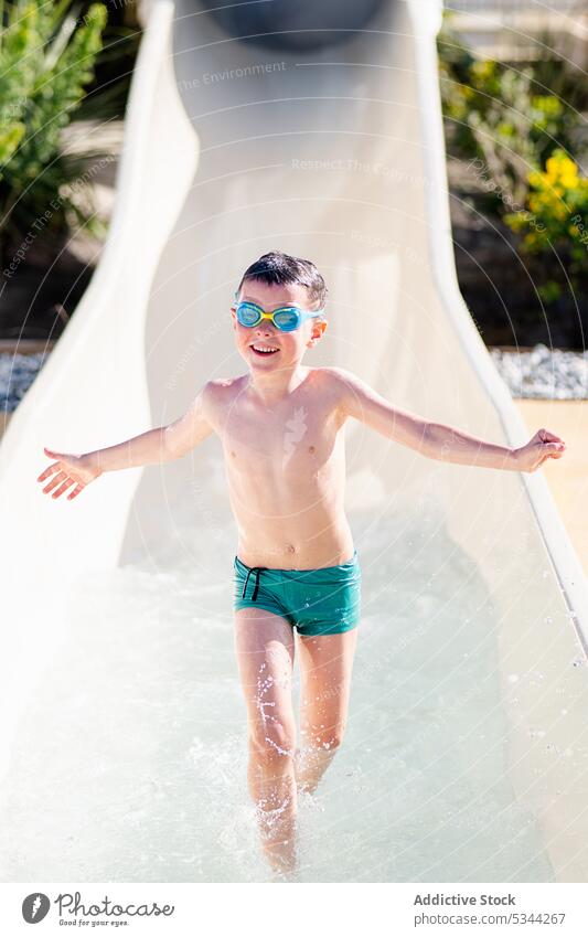 Glücklicher Junge läuft auf Wasserrutsche Daumen hoch gestikulieren Lächeln Resort Urlaub Sommer Badebekleidung heiter Schutzbrille Kind Feiertag genehmigen