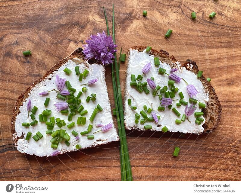 Eine Scheibe Brot mit Frischkäse, Schnittlauchröllchen und Schnittlauchblüten liegt auf einem Holzbrett Brotscheibe Belegtes Brot Appetit & Hunger Abendessen