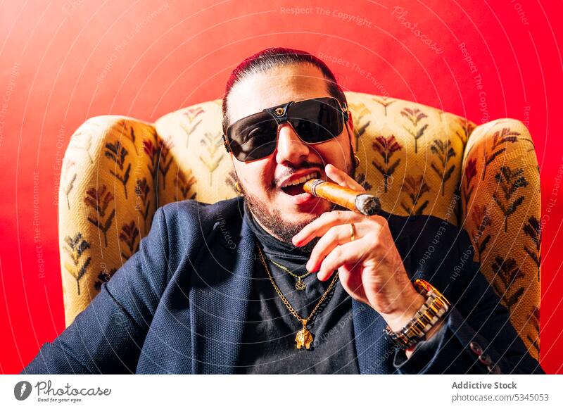 Glücklicher Mann mit Zigarre im Mund in einem Sessel in einem Studio Anzug Mode Raucherin Nikotin Süchtige Schlag männlich Armsessel heiter selbstbewusst