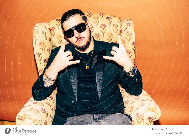 Ruhiger Mann zeigt Rockgesten im Sessel im Studio Stil Sonnenbrille Hipster gestikulieren Felsen ernst selbstbewusst Accessoire trendy männlich Mode Vollbart