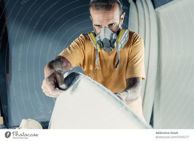 Fokussierter Mann mit Sandpapier auf dem Surfbrett polnisch Arbeit Schleifpapier Werkstatt professionell Mundschutz Meister behüten Beruf manuell Fähigkeit