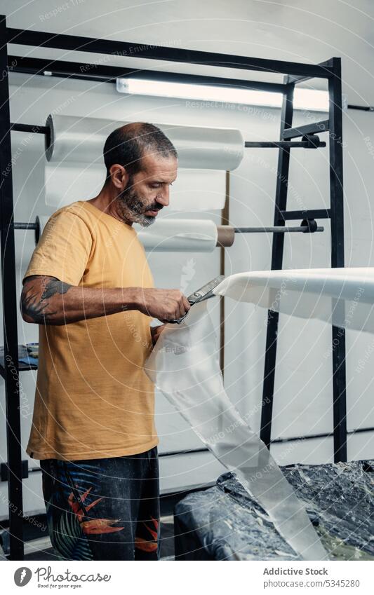 Fokussierter Mann schneidet Glasfasertuch auf Surfbrett Fiberglas geschnitten Schere messen Werkstatt professionell Arbeit Kunstgewerbler Instrument Werkzeug