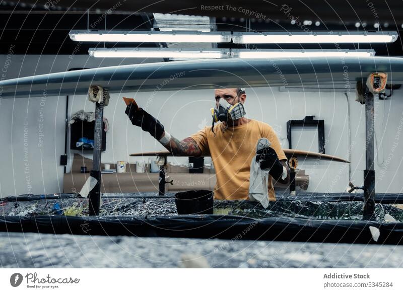 Fokussierter Mann, der einen Film mit einem Spachtel anbringt Surfbrett Atemschutzgerät behüten Werkstatt Meister befestigen kreieren Fiberglas sanft Schaber