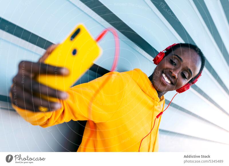 Von unten fröhliche ethnische Frau beim Videoanruf mit Smartphone und Kopfhörern Lächeln modern Glück Straße positiv heiter Apparatur Gerät urban Anschluss