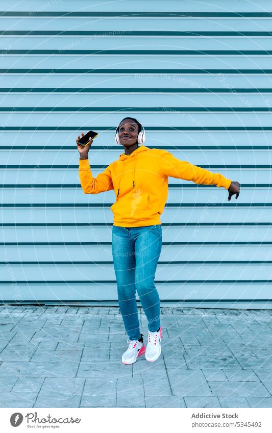 Fröhliche junge ethnische Frau in trendiger Freizeitkleidung und mit Kopfhörern tanzt auf der Straße mit Mobiltelefon Tanzen Musik Smartphone Wand Stil Lächeln