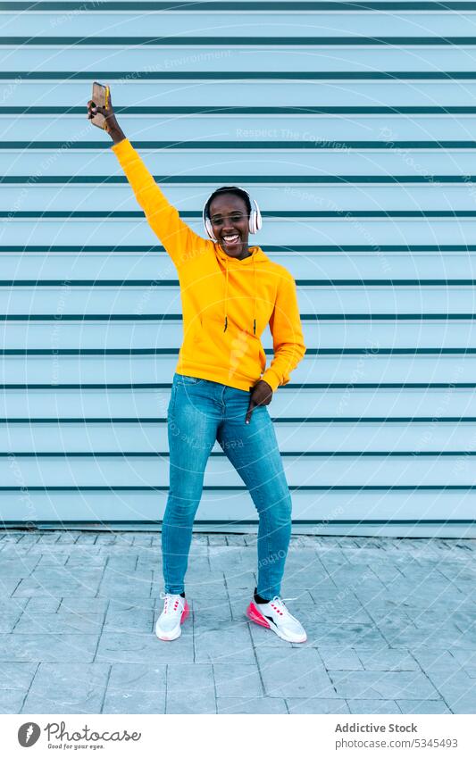 Fröhliche junge ethnische Frau in trendiger Freizeitkleidung und Kopfhörern tanzt auf der Straße mit Handy Tanzen Musik Smartphone Wand Stil Lächeln Glück Gerät