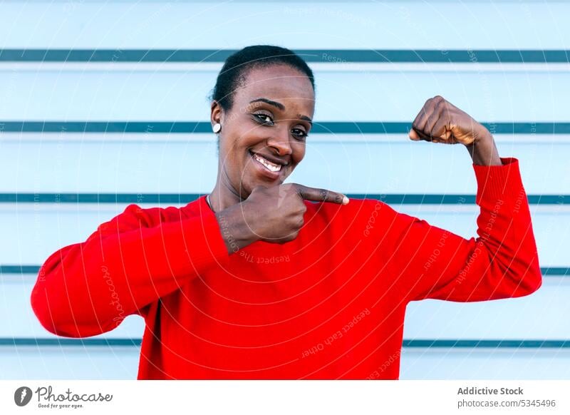Freudige Frau zeigt Muskeln auf der Straße Bizeps Punkt zeigen Lächeln Inhalt Porträt schwarz Afroamerikaner passen stark manifestieren gestikulieren expressiv
