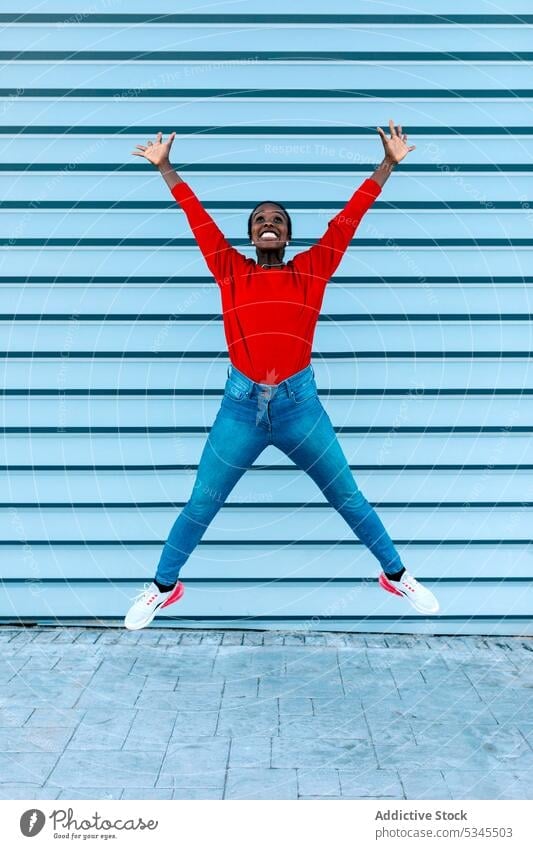 Aufgeregte Frau springt mit erhobenen Armen Model springen Sprung aufgeregt heiter Lächeln Großstadt Straße schwarz Afroamerikaner feiern Freude begeistert