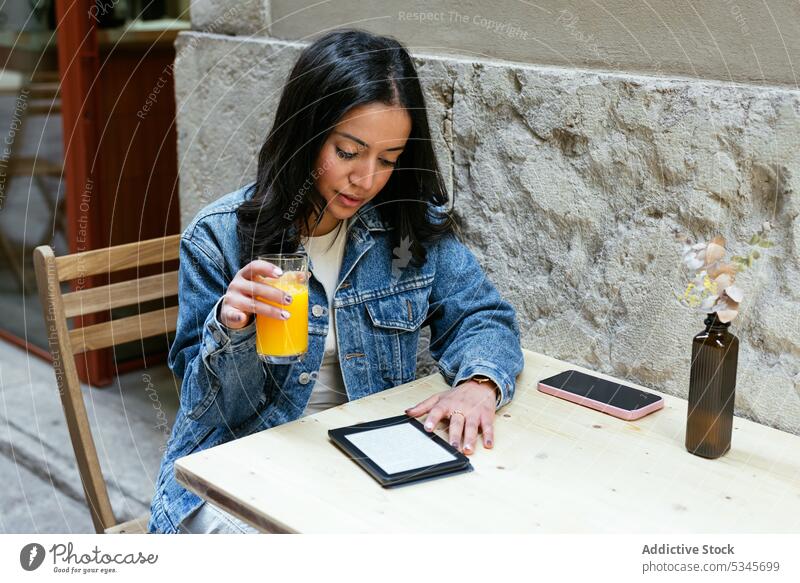 Konzentrierte ethnische Frau hält ein Glas Orangensaft in einem Straßencafé benutzend Tablette Saft trinken orange Internet online Smartphone Café Funktelefon