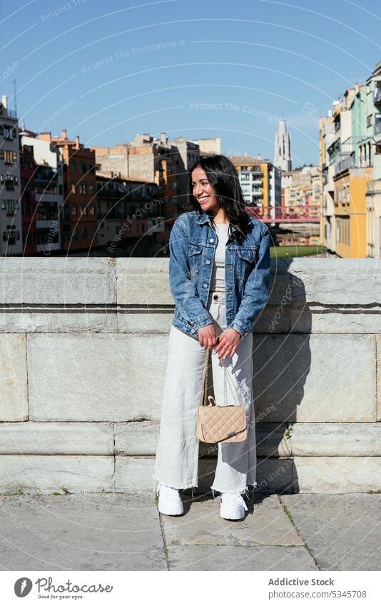Glückliche ethnische Frau steht in der Nähe der Mauer auf der Terrasse Lächeln Wand Handtasche Blauer Himmel Gebäude urban froh Stadtbild wolkenlos jung Sommer