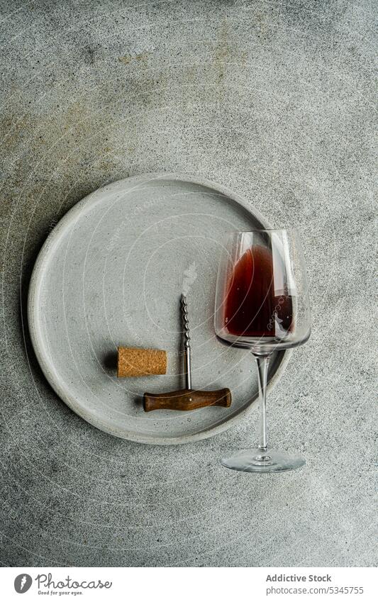 Hoher Blickwinkel auf ein Glas trockenen Rotweins in einem Glas, das auf einem Teller mit einem Korkenzieher und einem Korken auf einem Betonhintergrund liegt