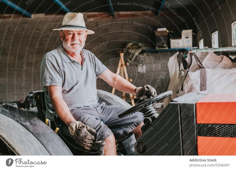 Älterer männlicher Landwirt sitzt auf einem Traktor in einer Garage Mann Porträt Senior Landschaft Ackerbau Maschine gealtert ländlich Fahrzeug schäbig sitzen