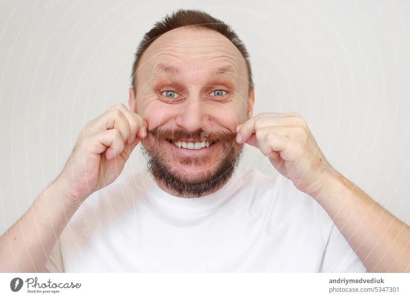 Porträt der glücklichen kaukasischen bärtigen Mann berührt seinen Schnurrbart in weißen lässigen T-Shirt lächelt und schaut in die Kamera auf weißem Hintergrund. Lifestyle und positive Emotionen Konzept. Art und glückliches Gesicht